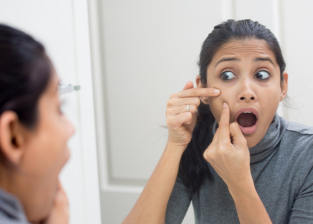 Young brown skin woman extracting acne in the mirror with a surprised/painful look on her face.